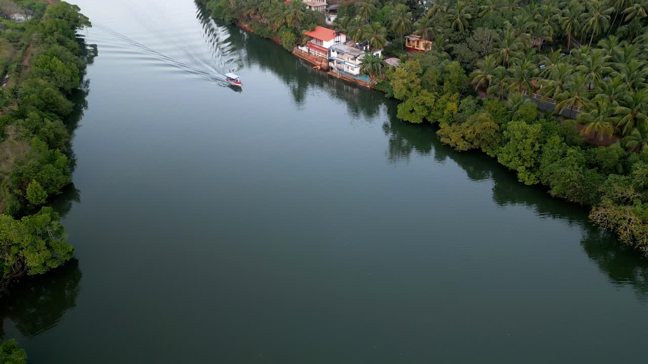 A top drone shot of a serene boat gliding on a tranquil Goan river India, leaving gentle ripples behind. The river is flanked by lush green trees, painting a peaceful, tropical paradise
