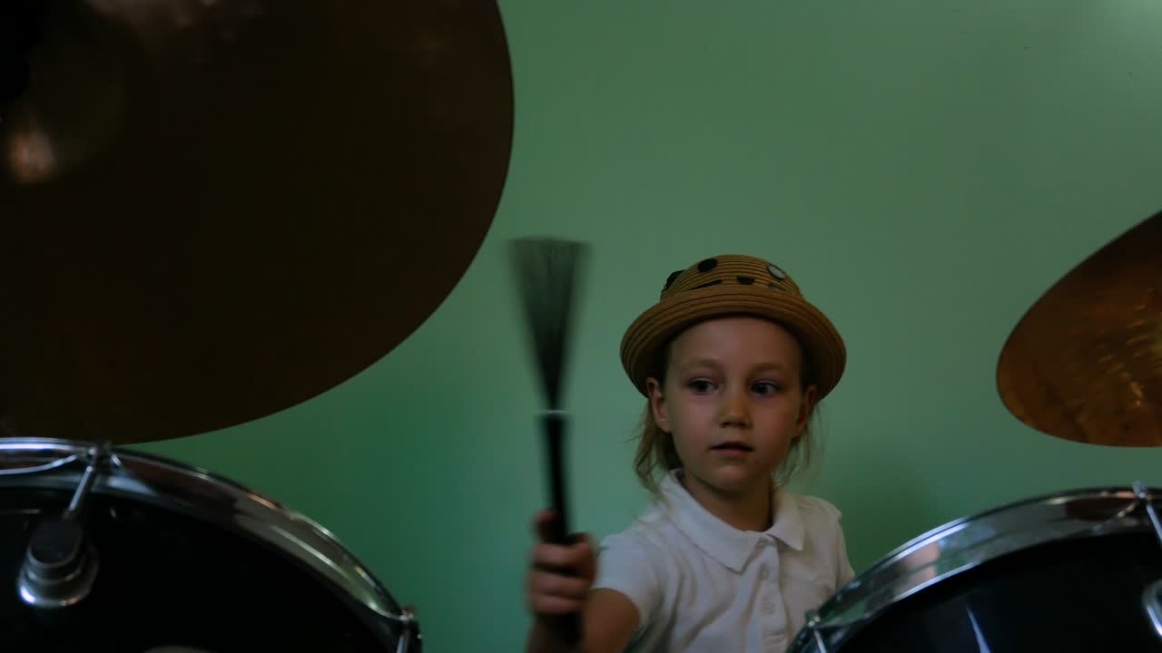 niño estudiando batería en la escuela de música. kit de batería negra. pequeño músico baterista con pinceles de batería entrenando en el fondo de la pared verde. niña rubia con sombrero jugando y sonriendo en la lección. 4k