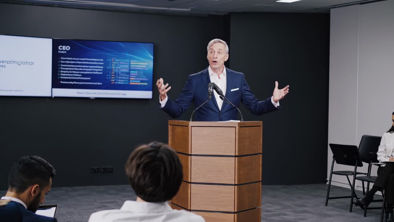 A video still of a business presentation, shot from a low angle, featuring a speaker at a podium