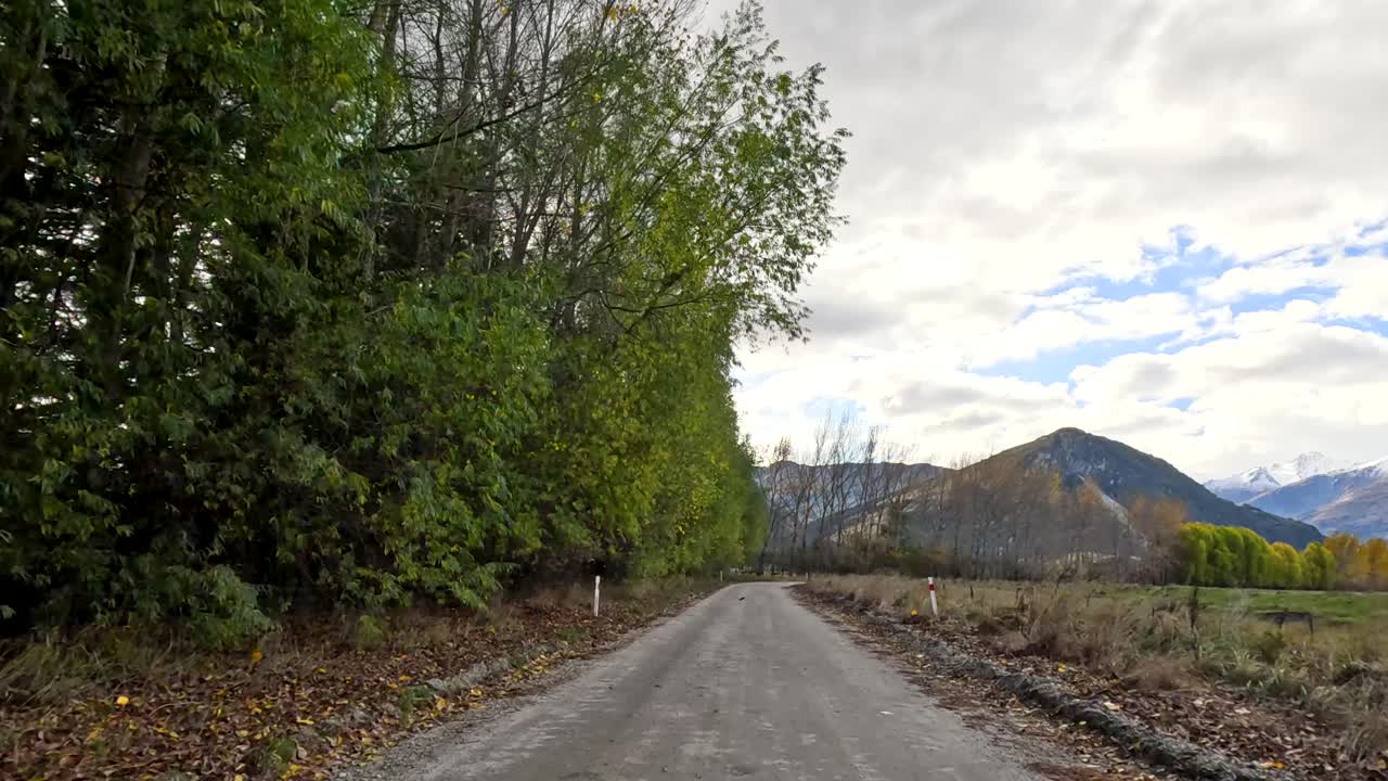 Vehicle moves along rural gravel road, passing dense trees, toward distant mountains under cloudy sky