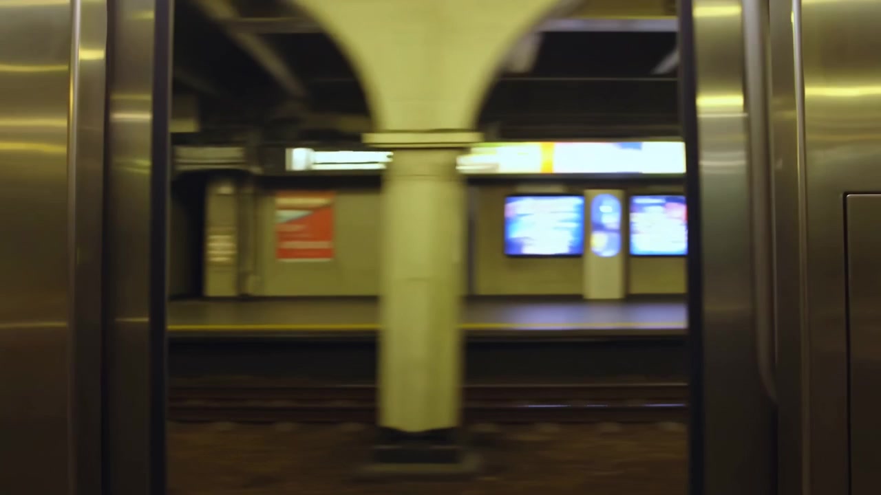 Prospettiva interna di una stazione della metropolitana con superfici metalliche e pilastri in cemento, atmosfera neutra, ambientazione di trasporto pubblico, contesto di vita urbana, illuminazione gialla e bianca, pulita.