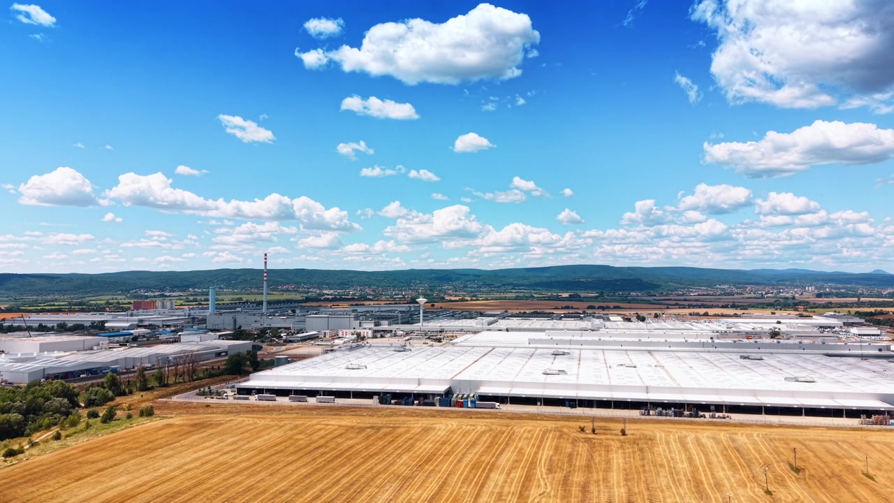 Ampia vista aerea di un enorme impianto di produzione. Un vasto edificio industriale con tetto bianco si trova adiacente a un campo agricolo secco sotto un cielo nuvoloso.