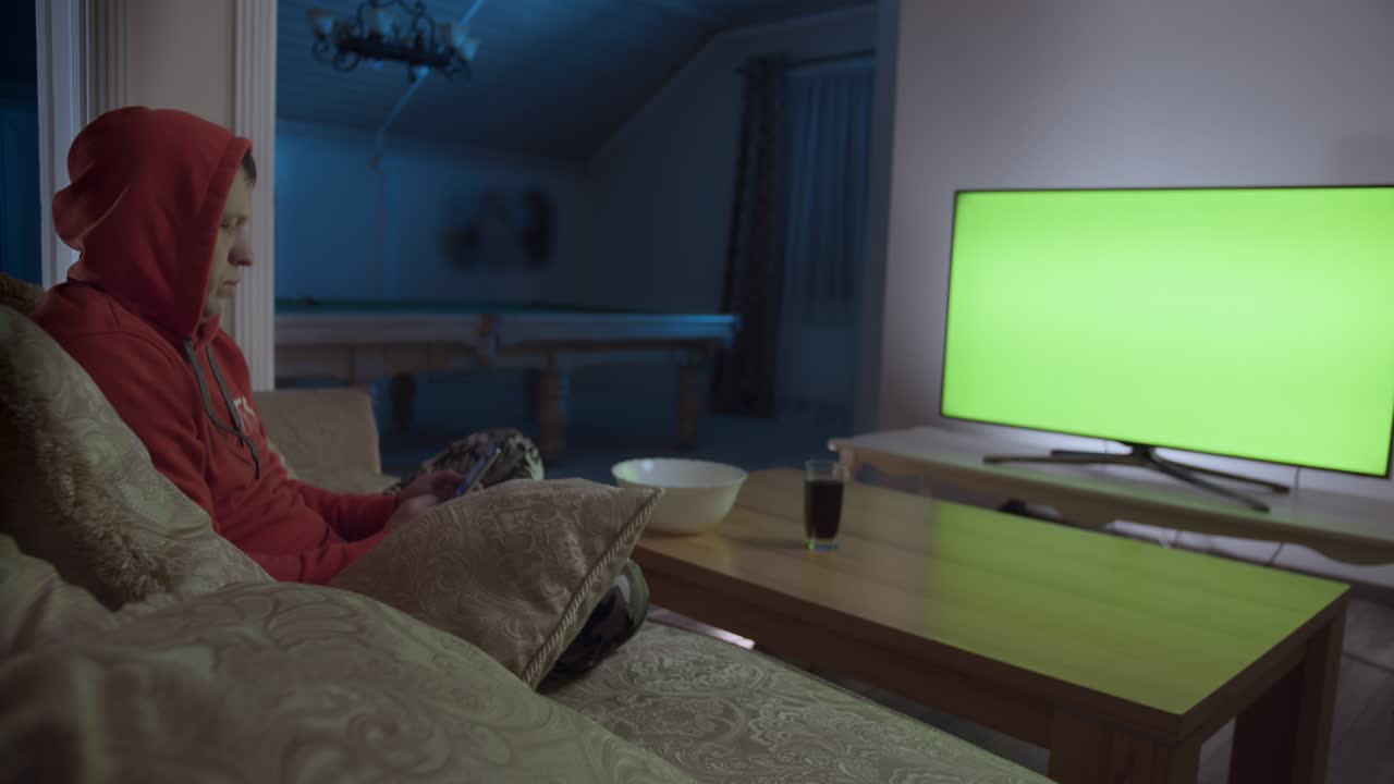 Uomo seduto su un divano con un telefono cellulare davanti a una TV con schermo verde.
