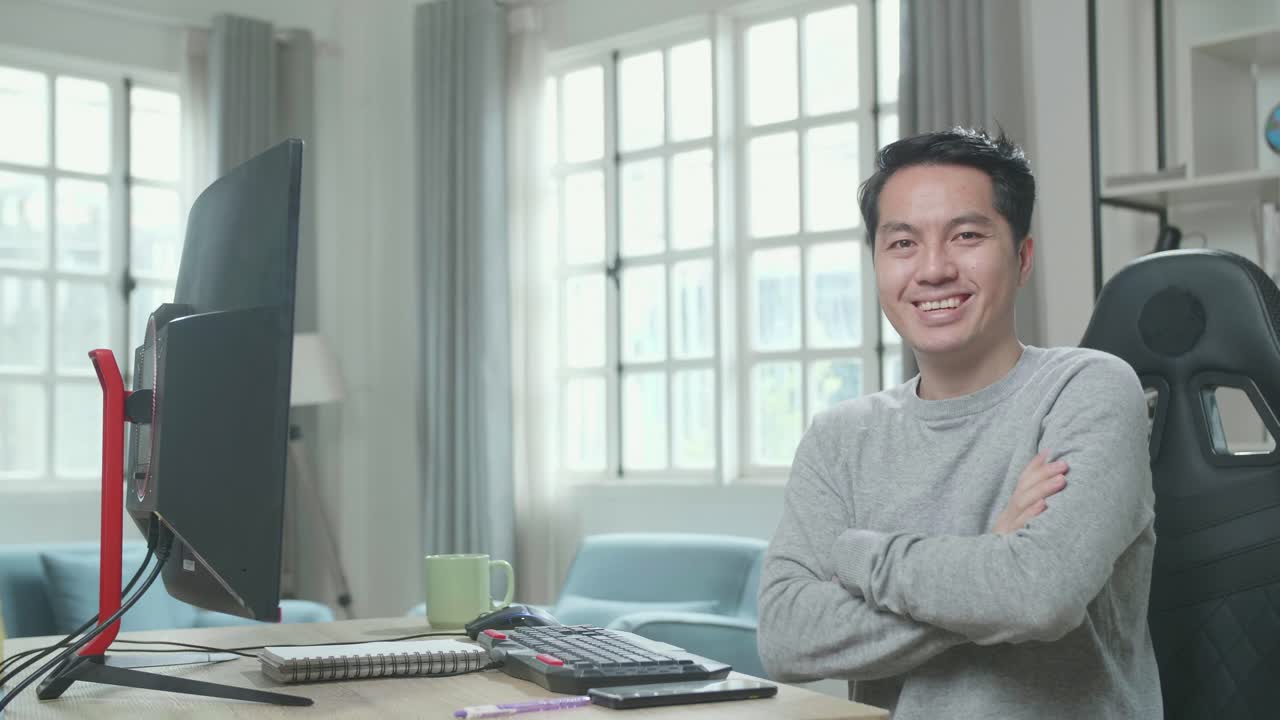 Asian Man In Long Sleeved T-Shirt Crossing His Arms And Smiling To Camera While Working With Desktop Computer At Home.