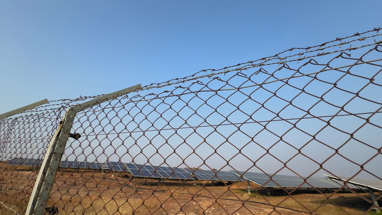 Panning view of a vast solar farm behind a chain-link fence under a clear blue sky, showing long rows of solar panels stretching across dry open land