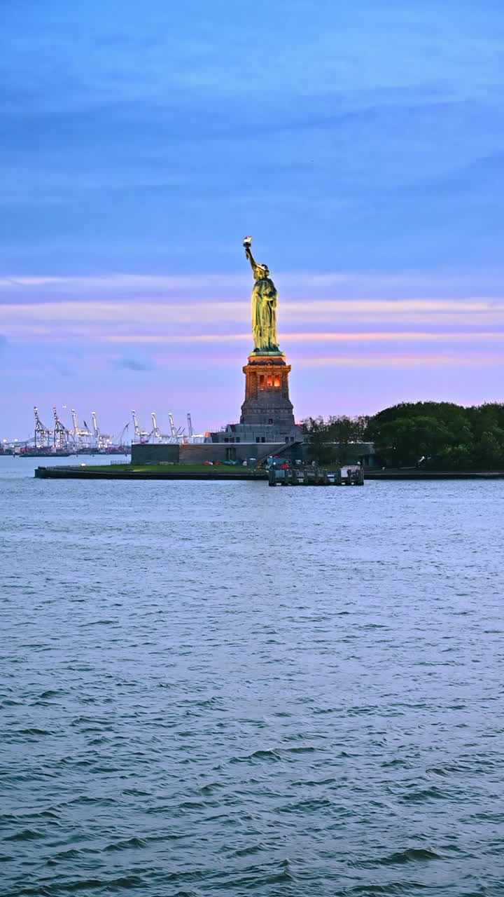 New York, AS, 8 Agustus 2025: Bepergian menyusuri Sungai Hudson saat matahari terbenam. Mendekati Patung Liberty dengan pencahayaan malam. Memuat derek di pelabuhan sebagai latar belakang. Video vertikal