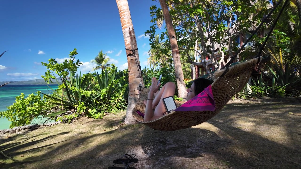 Mujer relajándose en hamaca leyendo un libro electrónico junto al mar tropical turquesa