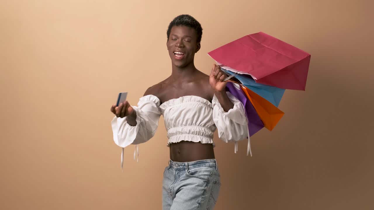Happy transgender person showing a credit card holding shopping bags over beige background