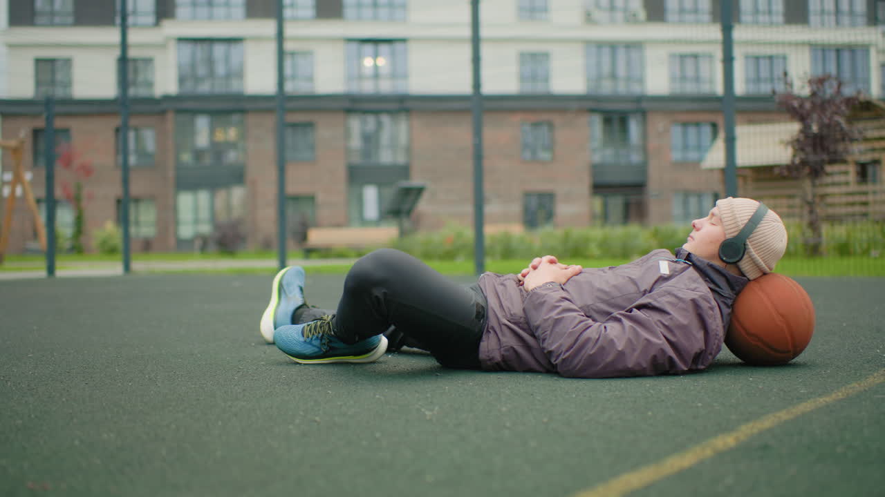 Eine ältere Frau liegt mit einem Ball als Kopfstütze auf einem Basketballplatz, trägt Kopfhörer und eine violette Jacke, Turnschuhe sind sichtbar, Mehrfamilienhäuser und ein Zaun im Hintergrund, trübes Licht, reflektierende, ruhige Stimmung