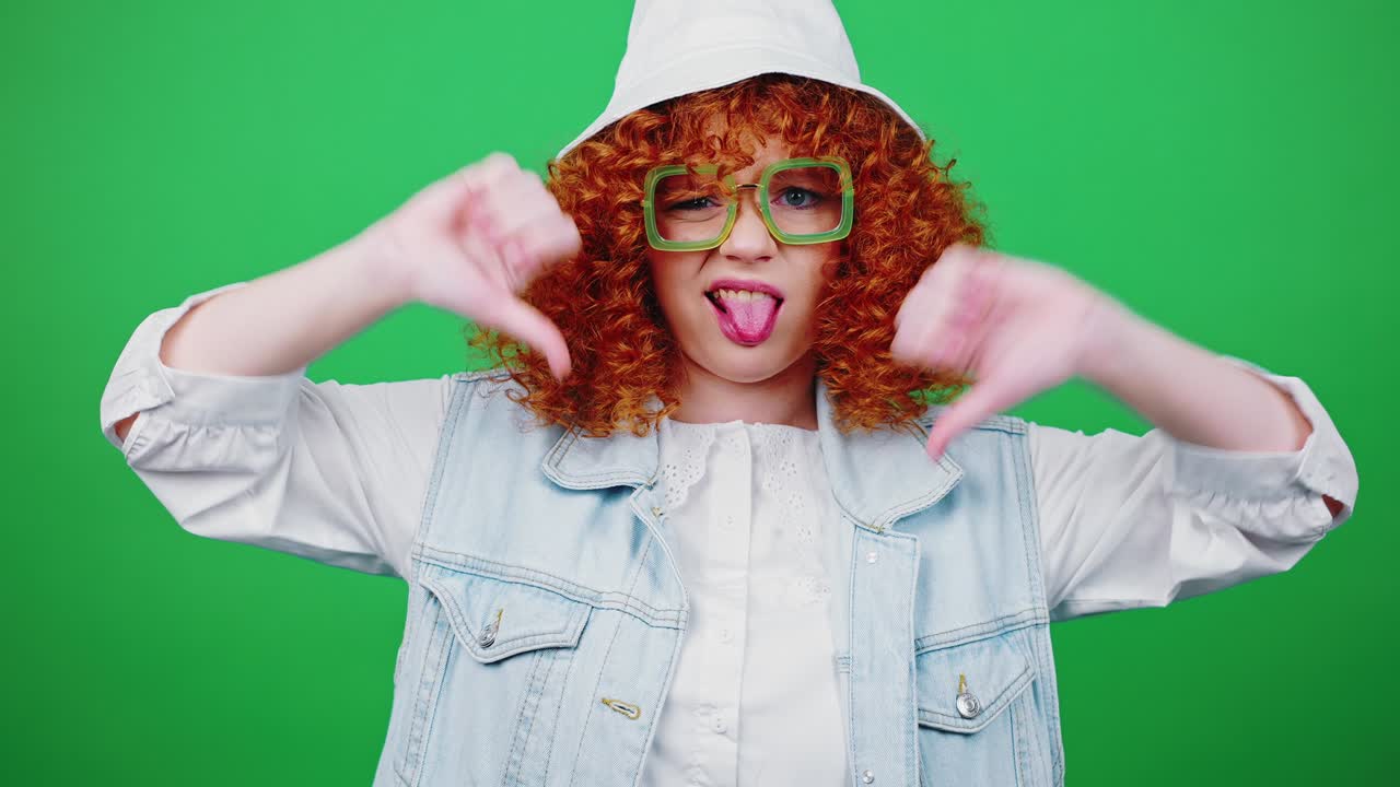 Dislike concept. Young redhead curly woman showing thumbs down gesture, expressing denial, green chroma key background