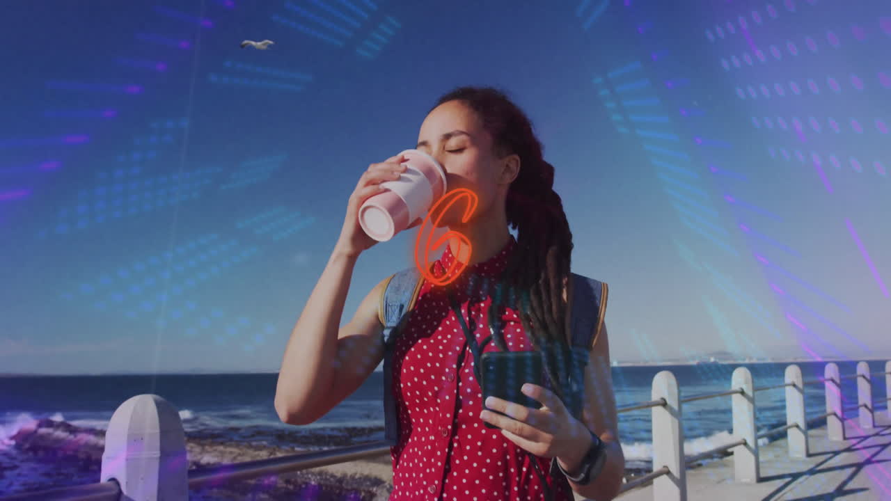 Woman holding smartphone and coffee cup on seaside promenade, showcasing technology light overlays