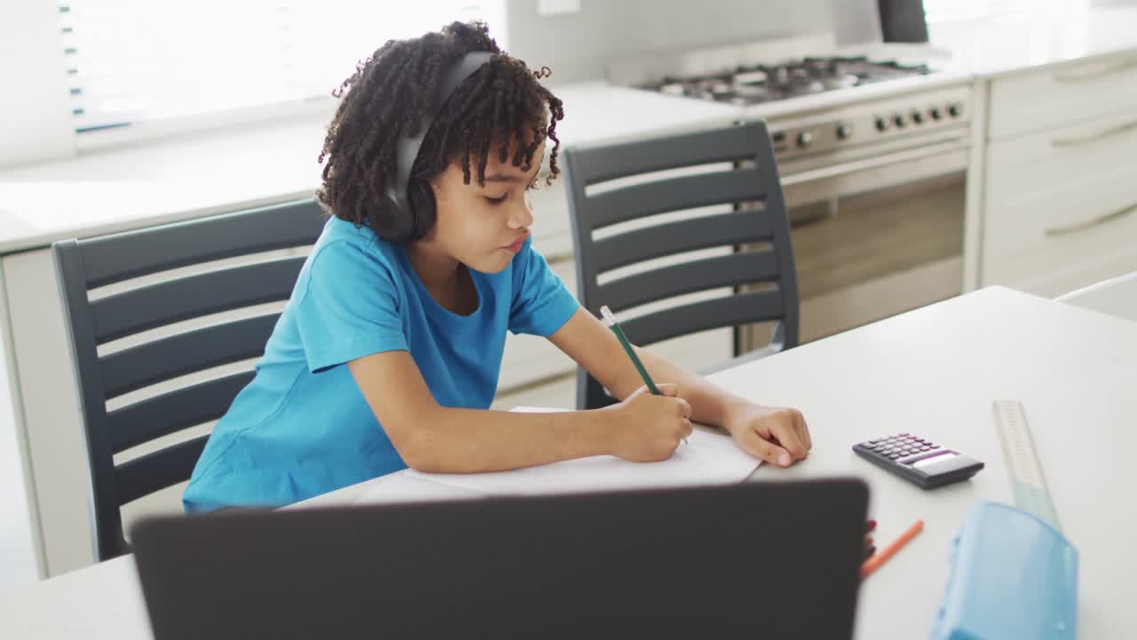feliz muchacho biracial sentado en la mesa en la cocina usando computadora portátil y haciendo la tarea