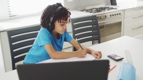 Happy-biracial-boy-sitting-at-table-in-kitchen-using-laptop-and-doing-homework