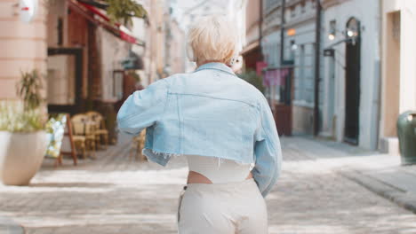 carefree young woman in glasses listening music via wireless headphones while walking on city street