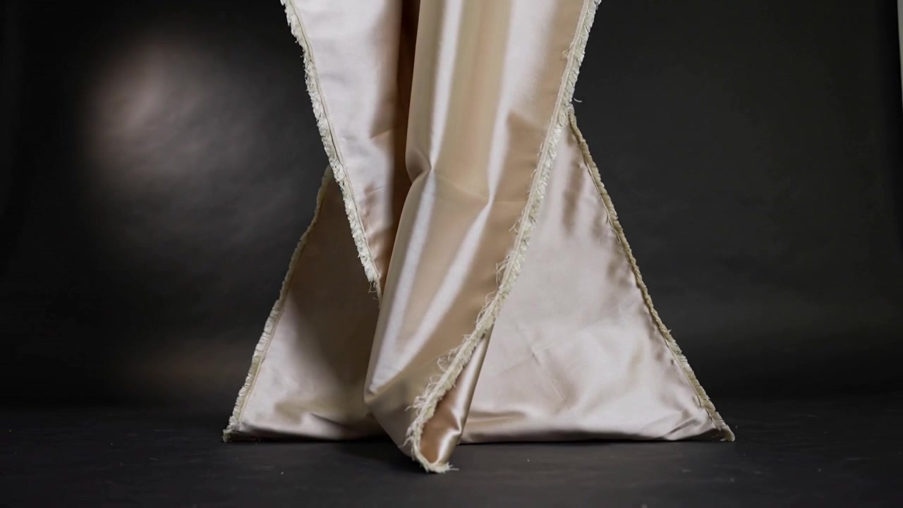Tessuto di raso beige che si srotola su un pavimento scuro con sfondo nero, adagiandosi simmetricamente