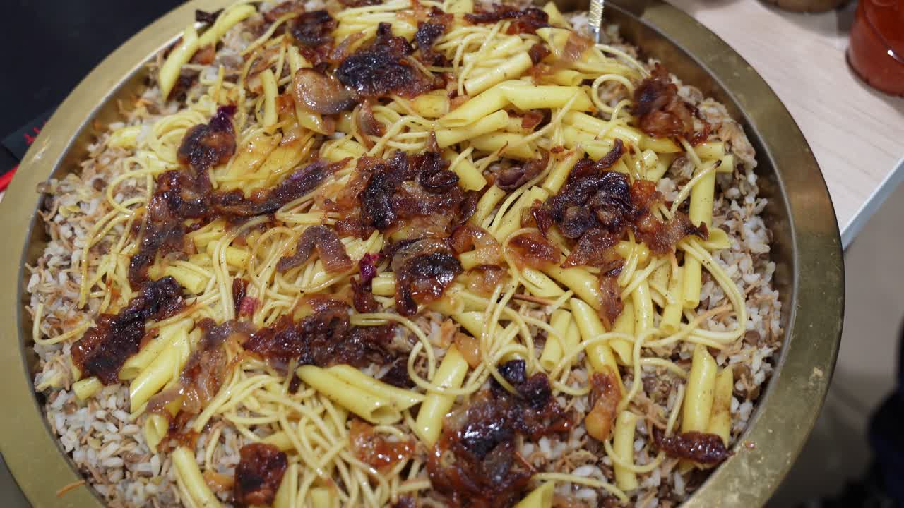 室内で、温かい光の下、テーブルの上で撮影された、ご飯、パスタ、レンズ豆、クリスピーオニオンを添えたエジプトのコシャリが大きなトレイから盛り付けられているクローズアップ