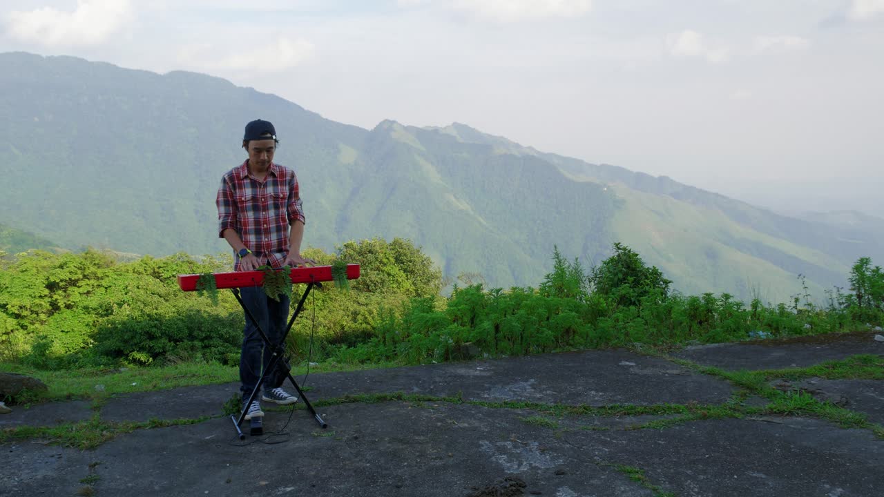 una grabación estacionaria de un hombre de la línea izquierda de la regla de los tercios, tocando su teclado musical electrónico con vistas a la montaña de fondo