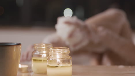 vista de cerca de una mesa con velas e incienso, en el fondo una mujer borrosa tomando un baño mientras se frota las piernas con jabón en el fondo