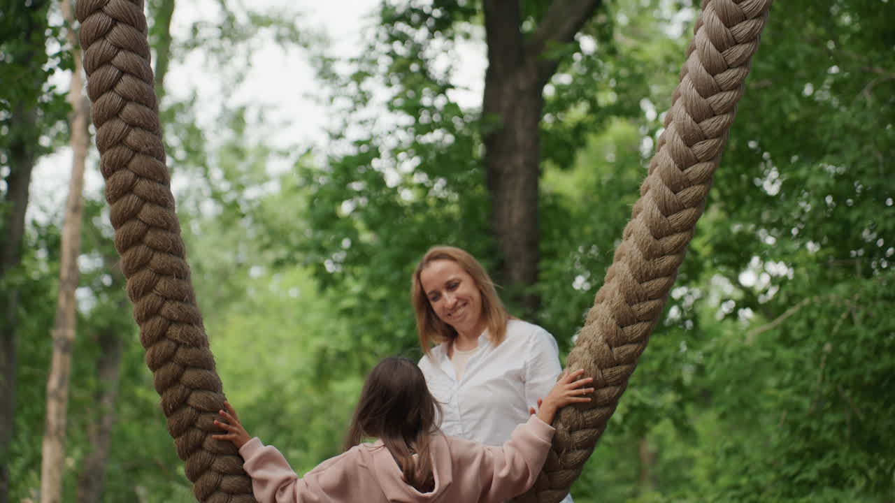 Hvid mor og datter nær gynge i skovlignende park, datter griber tyk flettet reb, mens mor ser på med et støttende smil, legende vejledning, roligt opsyn, ægte familieaktivitet, sommer.