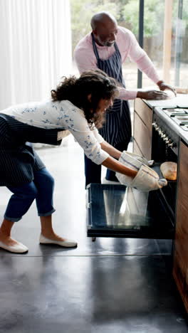vertical video of happy diverse mature couple baking at home, taking bread from oven, slow motion