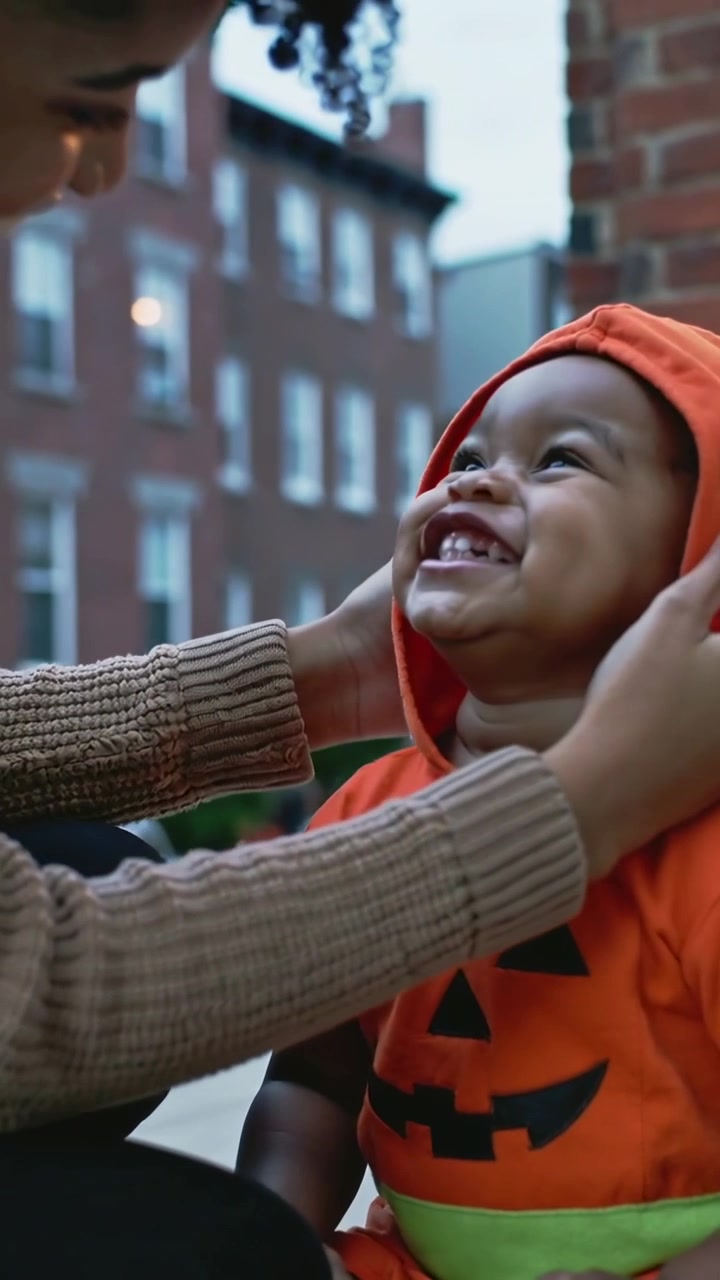 Video verticale: Mamma in maglia beige che si china mettendo un cappuccio a forma di zucca a un bambino piccolo sul portico, sistemando il cappuccio.