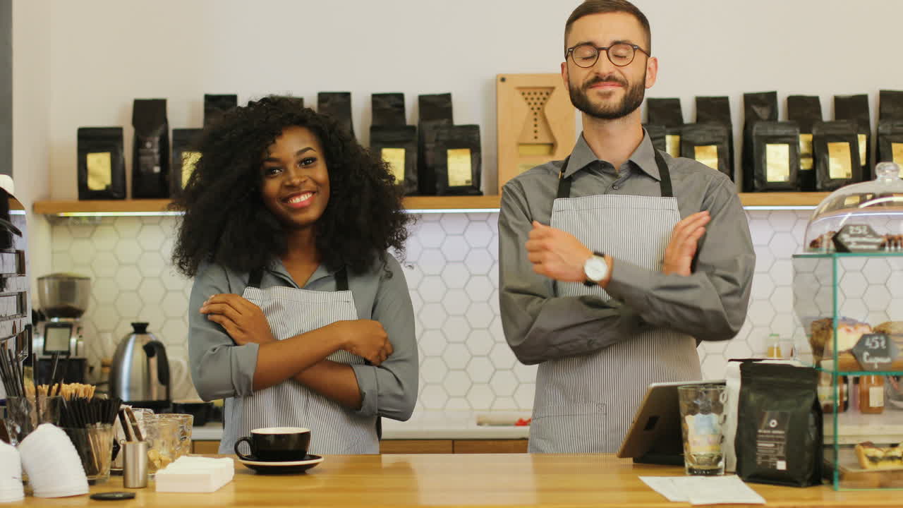 African american female barista and her male coworker smiling and looking at the camera with crossed arms behind the bar of coffee shop