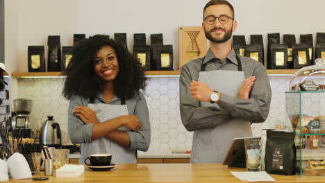 african american female barista and her male coworker smiling and looking at the camera with crossed arms behind the bar of coffee shop