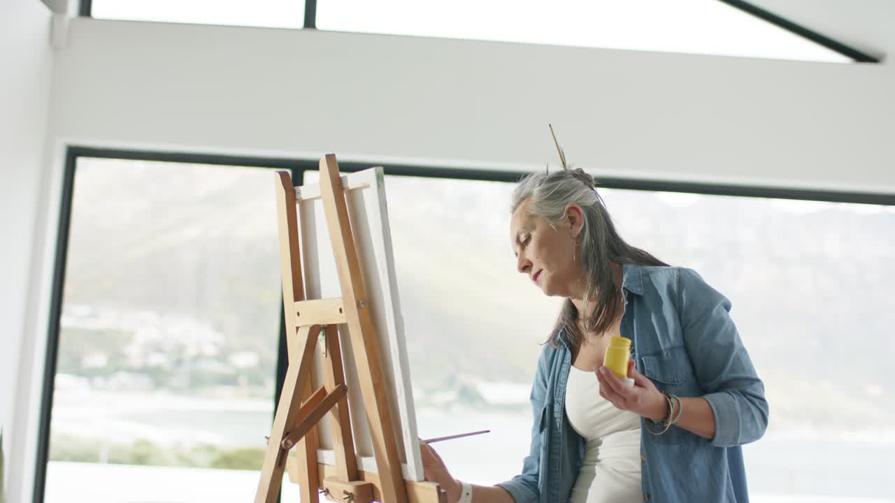 Moden kvinde holder gul malingkrukke, maler strøg på lærred i atelier ved vindue og skaber kunst.