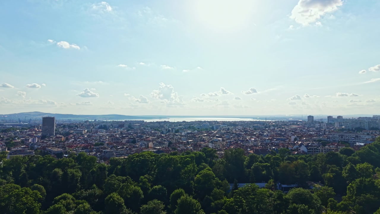 Blick auf eine Stadt mit Gebäuden, Bäumen und Wasser an einem sonnigen Tag, der Aktivität und Leben in der Gegend zeigt, mit klarem Himmel und vorbeitreibenden Wolken.
