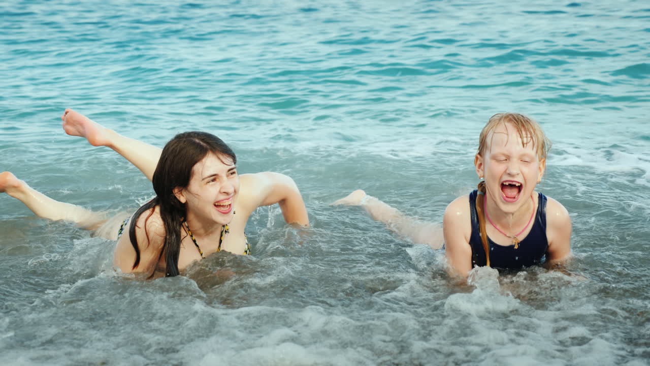 소녀와 함께 있는 젊은 활동적인 어머니가 재주 넘기며 웃고 있는 해변에서 바다 파도 위에서 즐겁게 놀고 있습니다.