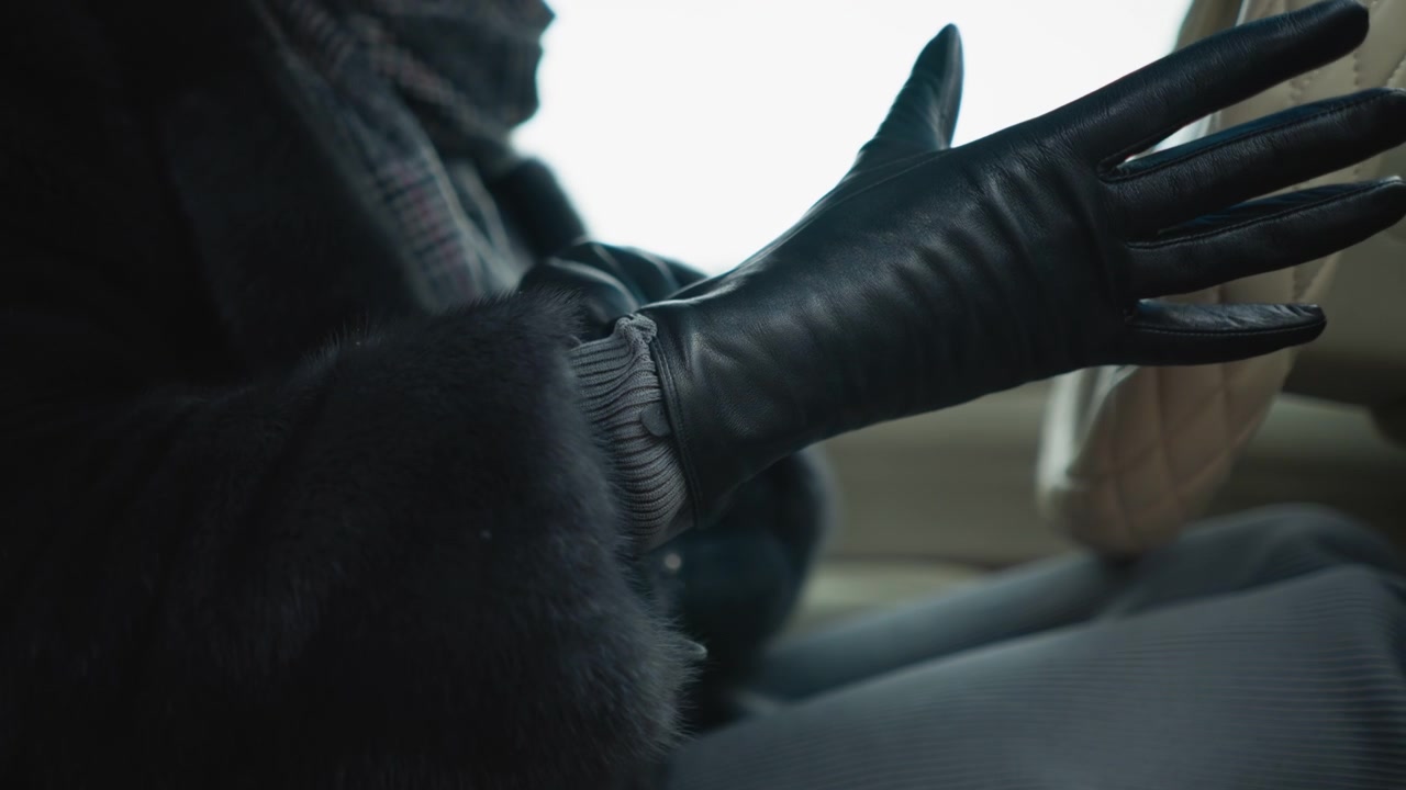 Close up of woman in black leather gloves adjusting them while seated inside car. focus remains on her hands and texture of glove material with soft lighting highlighting details. Background