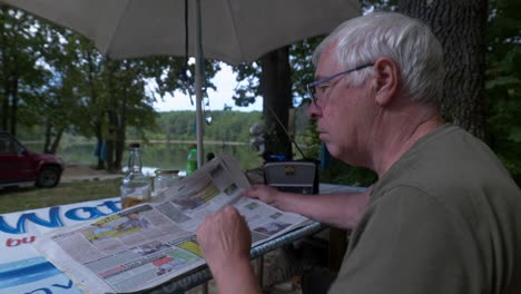 grey haired man sits outdoors in woodland scene reading local newspaper