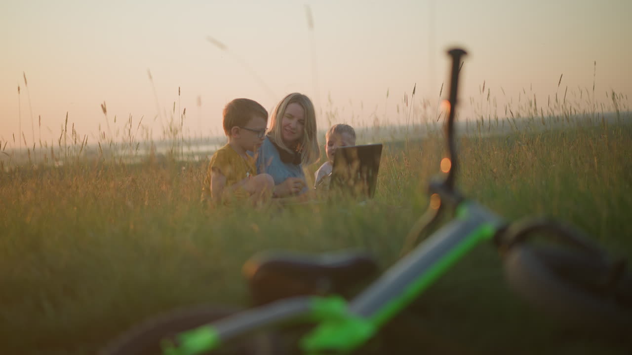 A mother in a blue gown shares a moment with her two sons, chatting happily while seated on a grassy field. A blurred bicycle lies nearby, enhancing the scene's relaxed, family-focused atmosphere