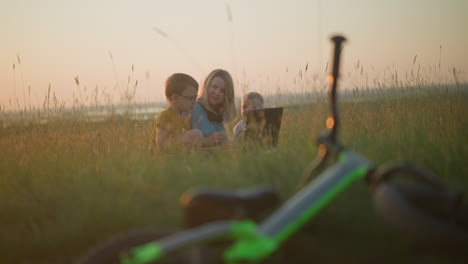 a mother in a blue gown shares a moment with her two sons, chatting happily while seated on a grassy field. a blurred bicycle lies nearby, enhancing the scene's relaxed, family-focused atmosphere
