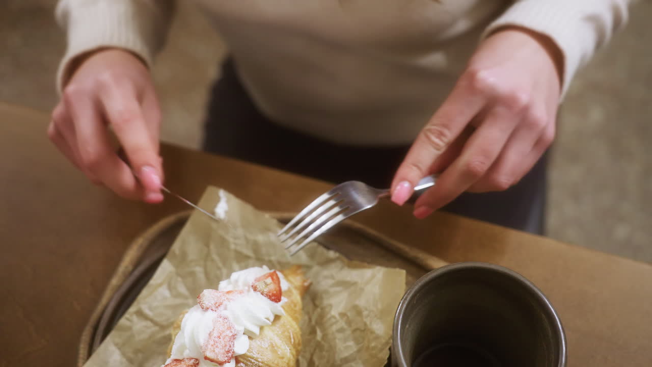 Close-up shot of female hand unwrapping fork in cozy cafe. The scene captures a croissant with whipped cream and a coffee cup on the table. Relaxed atmosphere with soft background lighting