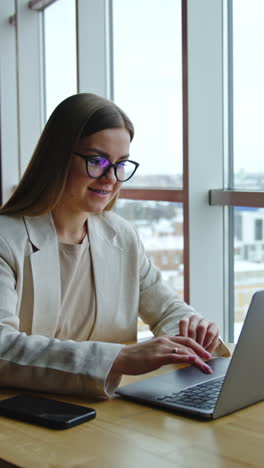 smiling business lady sitting at the desk and working at laptop. attractive woman at light office workplace. panoramic windows with cityscape at backdrop. vertical video