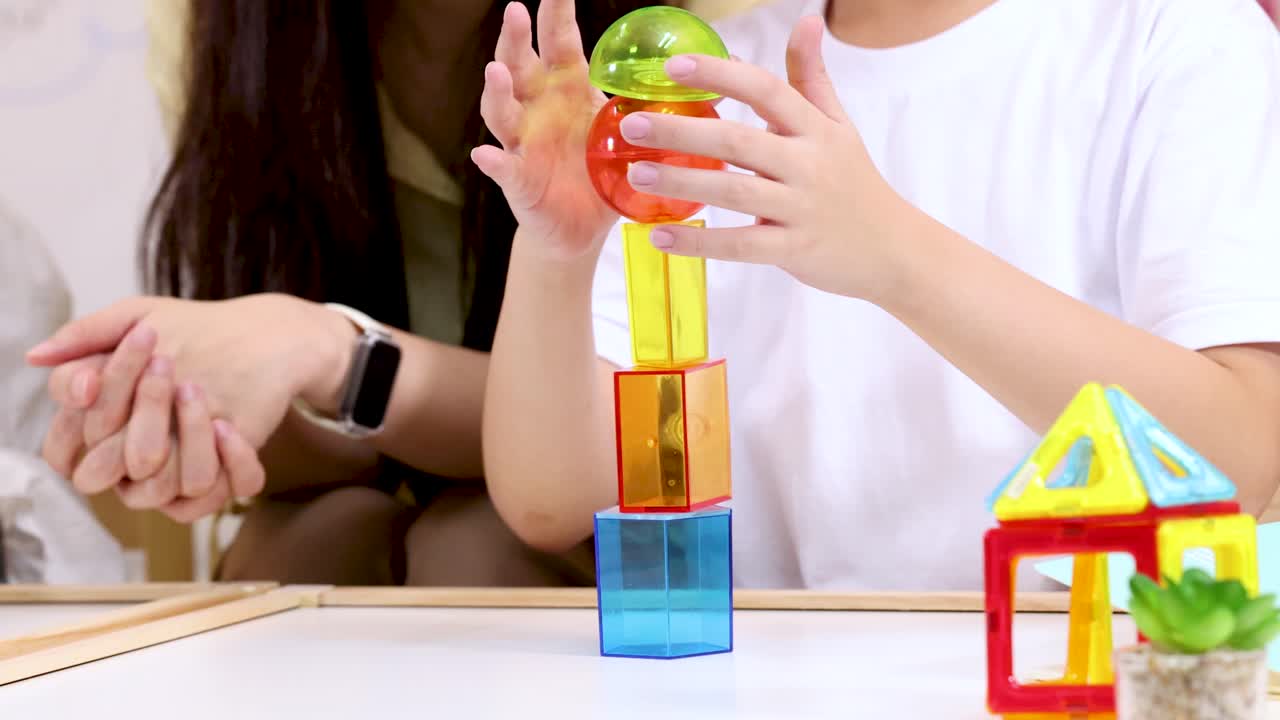 Un niño apila bloques de plástico de colores en un aula luminosa, guiado por la maestra, con tomas de primer plano estables.