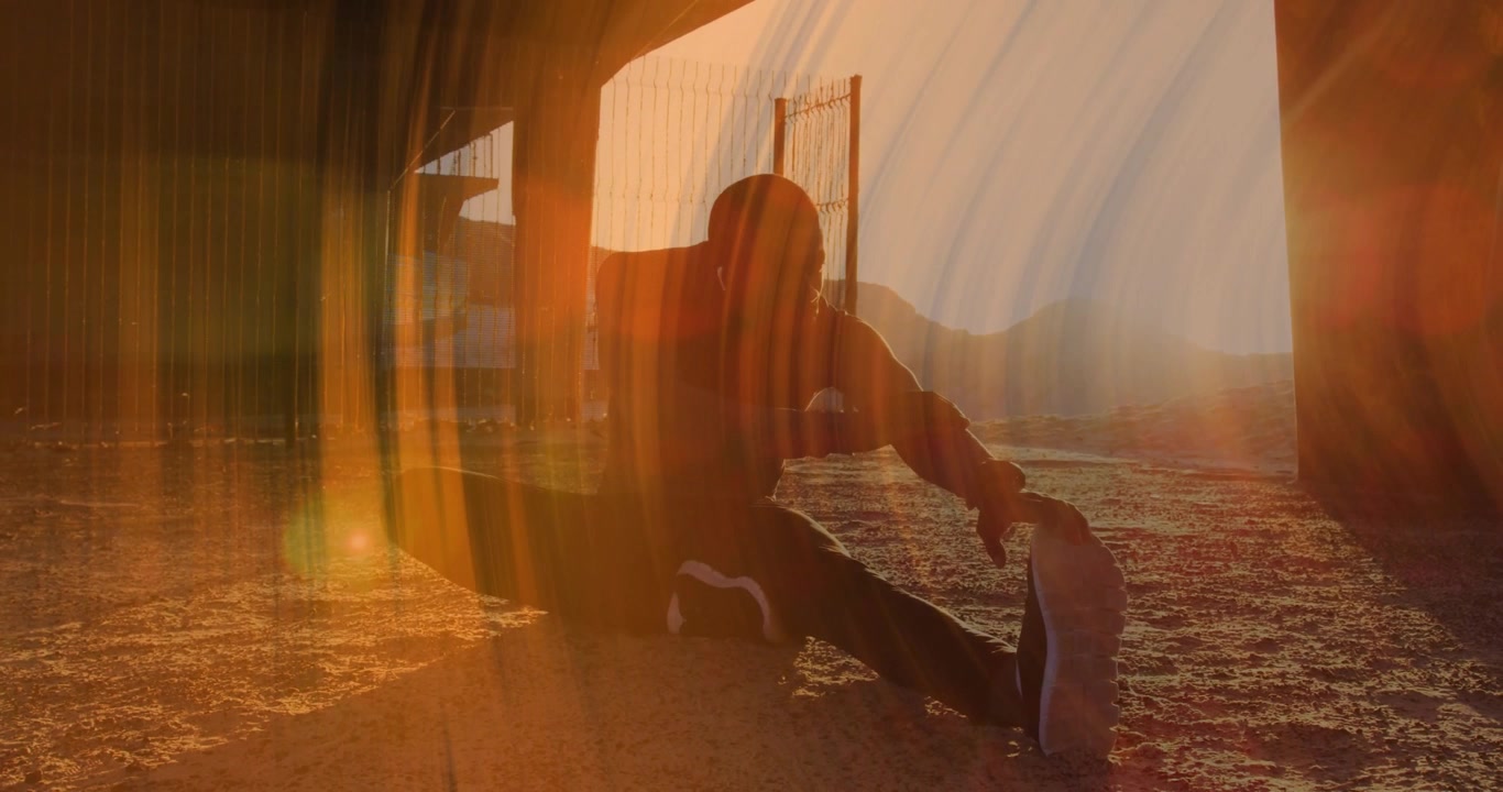 Uomo che sposta il peso sotto una tettoia ondulata, iniziando lo stretching dei femorali durante il riscaldamento al tramonto.