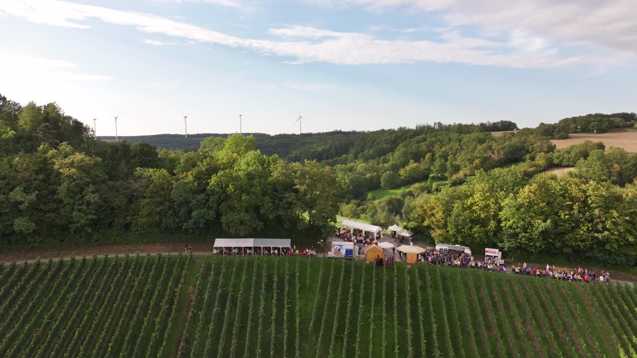 Aerial shot of a lively wine festival taking place during the afternoon within a vineyard on top of a forest mountain
