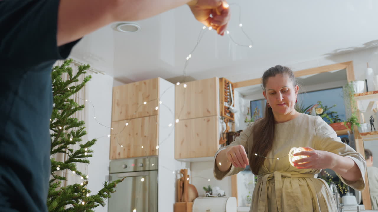 Husband assisting wife with Christmas lights, wife in gown neatly folding light strands, cozy home with Christmas tree in the background, festive holiday preparation