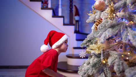 baby boy wearing red t-shirt and santa cap stands indoors. lovely kid decorates christmas tree.