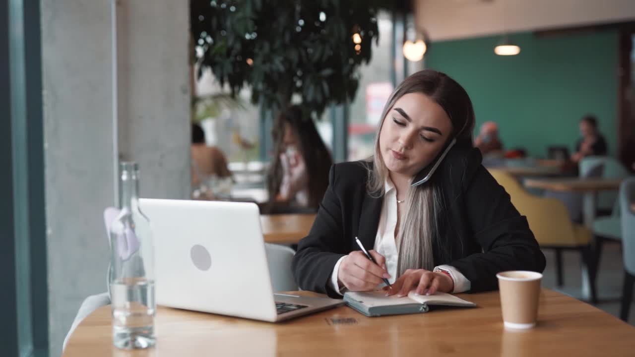 joven, hermosa mujer sentada en un café, vestida con ropa de negocios, ocupada hablando por teléfono, tomando notas en su cuaderno, y trabajando en su portátil