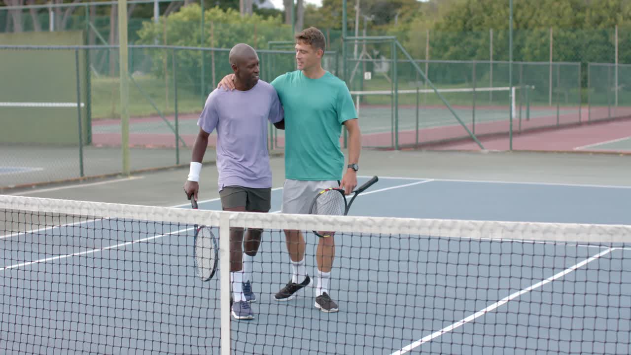 Portrait of two happy diverse male friends embracing on outdoor tennis court after game, slow motion