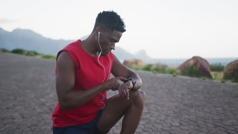 african american man wearing earphones using fitness band while sitting on the road