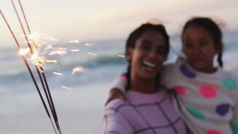 Portrait-of-happy-hispanic-mother-and-daughter-playing-with-sparklers-on-beach-at-sunset