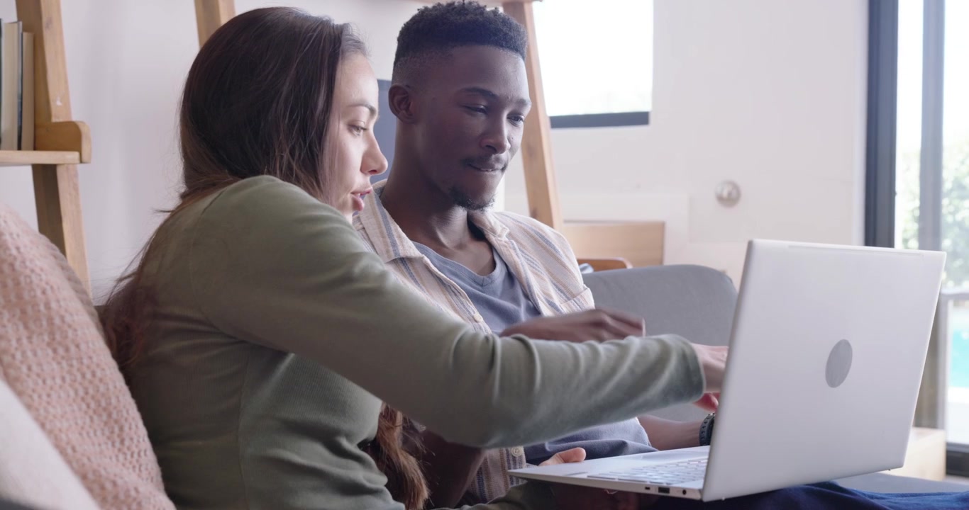 Diverse couple sitting on couch positioning silver laptop, typing and pointing while reviewing work