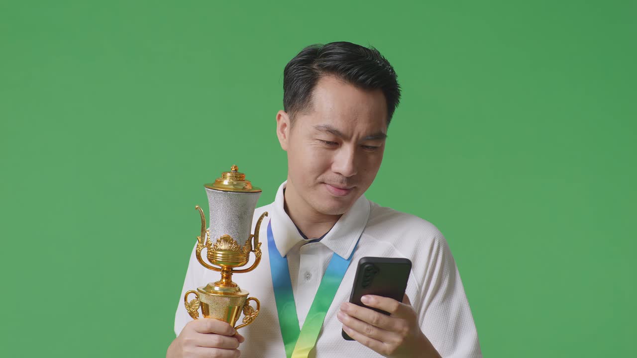 골드 트로피를 들고 스튜디오에서 녹색 화면 배경에 스마트폰을 사용하는 금메달을 가진 아시아 남자의 클로즈업