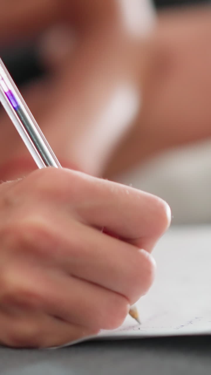 close up of young person writing with ballpoint pen while seated on sofa with slightly blurred background showing legs crossed and casual home environment