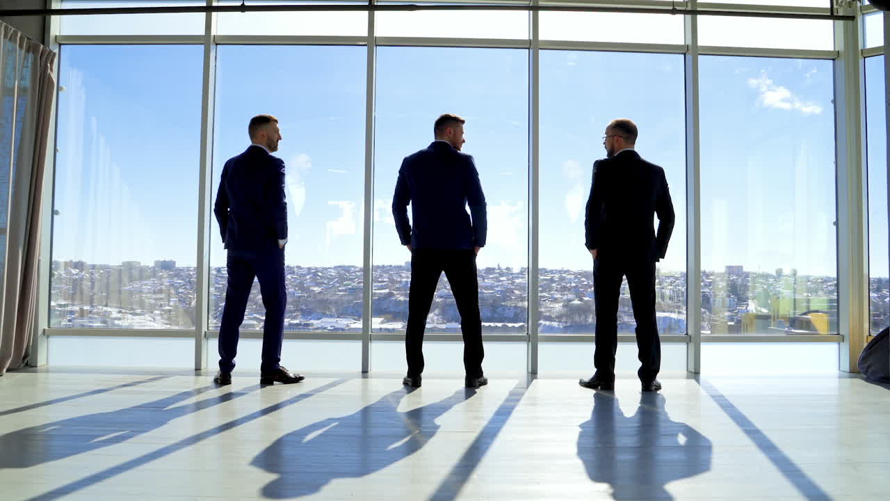 Business people in office on city landscape through window. Three men in elegant suits standing by the window and talking in sunny daytime. Back view.