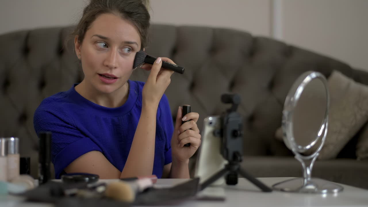 Woman in a blue T-shirt filming herself while doing a make-up tutorial at home